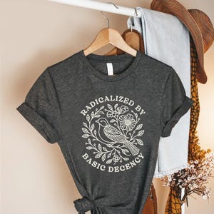 May include: Dark gray t-shirt with rolled sleeves, featuring a white graphic of a bird, flowers, and leaves, encircled by the text "RADICALIZED BY BASIC DECENCY". The shirt is hanging on a wooden hanger.