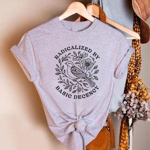 May include: A light gray t-shirt featuring a black graphic of a bird perched on a branch, surrounded by leaves and flowers. The text "RADICALIZED BY BASIC DECENCY" is arched around the design. The shirt is knotted at the waist.