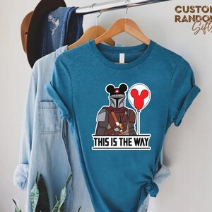 Puede incluir: Camiseta de color turquesa con un dise&ntilde;o de personaje mandaloriano, con una cabeza de Mickey Mouse y un globo rojo. La frase "THIS IS THE WAY" est&aacute; impresa debajo. La camiseta est&aacute; colgada en una percha de madera.