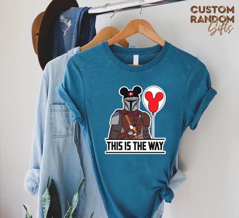 Puede incluir: Camiseta de color turquesa con un dise&ntilde;o de personaje mandaloriano, con una cabeza de Mickey Mouse y un globo rojo. La frase "THIS IS THE WAY" est&aacute; impresa debajo. La camiseta est&aacute; colgada en una percha de madera.