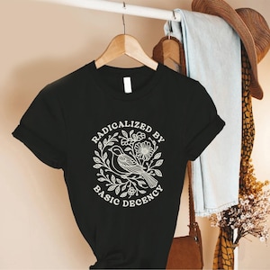 May include: Black t-shirt with the text "RADICALIZED BY BASIC DECENCY" in a white circular design featuring a bird, flowers, and leaves. The shirt is hanging on a wooden hanger.