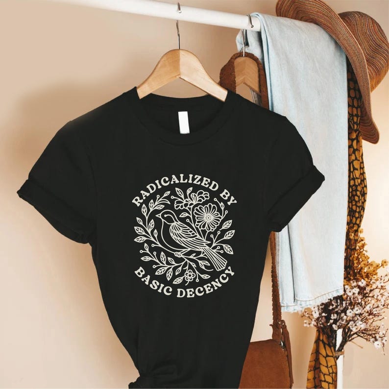 May include: Black t-shirt with the text "RADICALIZED BY BASIC DECENCY" in a white circular design featuring a bird, flowers, and leaves. The shirt is hanging on a wooden hanger.