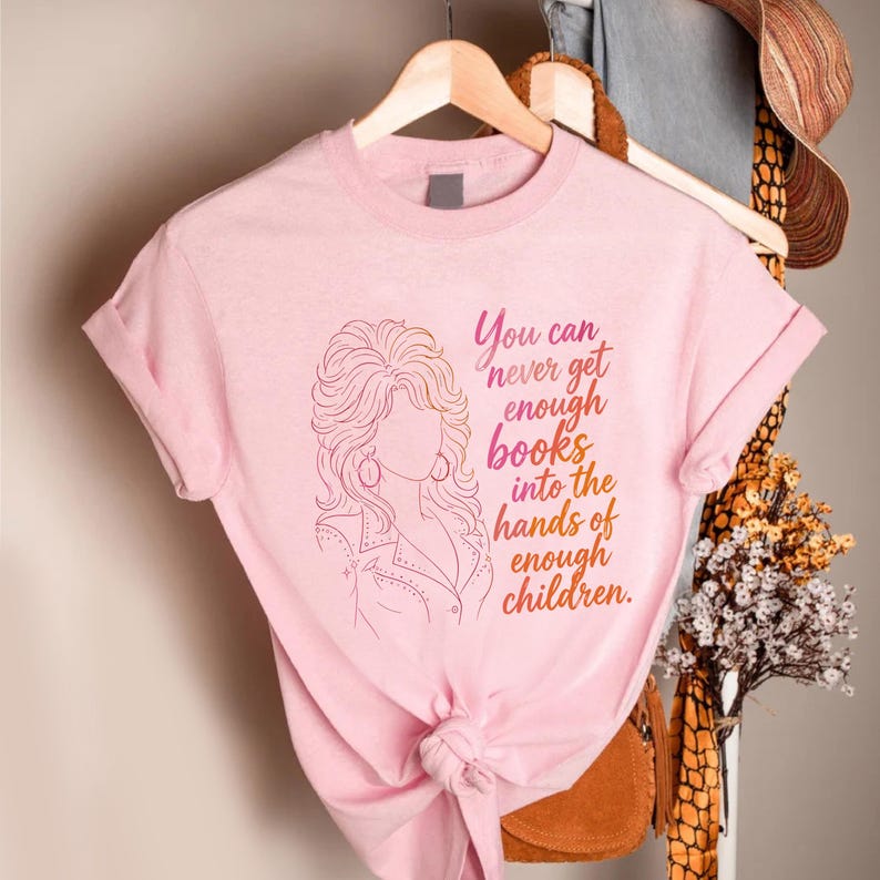 May include: A light pink t-shirt featuring a line art illustration of a person with voluminous hair and the text, You can never get enough books into the hands of enough children, printed in a gradient of pink, orange, and yellow hues.