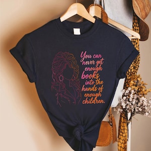 May include: A dark navy blue short-sleeve t-shirt featuring a pink and orange gradient line art illustration of a woman with voluminous hair and the text "You can never get enough books into the hands of enough children" in a matching gradient font.