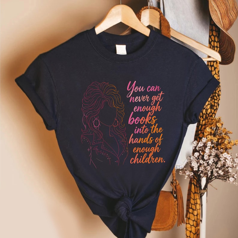 May include: A dark navy blue short-sleeve t-shirt featuring a pink and orange gradient line art illustration of a woman with voluminous hair and the text "You can never get enough books into the hands of enough children" in a matching gradient font.