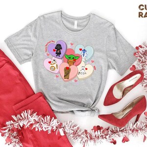 May include: A light gray t-shirt featuring a design of Star Wars characters on heart-shaped candies. The shirt is paired with red pants and red high heels. The text "Happy Valentine's Day" is on the shirt.