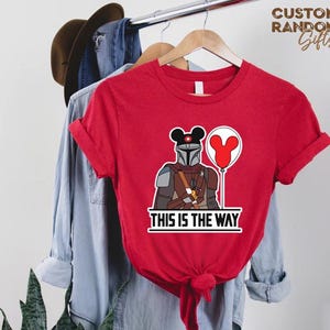 Puede incluir: Camiseta roja con un dise&ntilde;o del personaje de Mandalorian con orejas de Mickey Mouse y un globo. El texto "THIS IS THE WAY" est&aacute; impreso debajo. La camiseta est&aacute; colgada en una percha de madera.