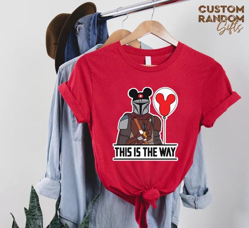 Puede incluir: Camiseta roja con un dise&ntilde;o del personaje de Mandalorian con orejas de Mickey Mouse y un globo. El texto "THIS IS THE WAY" est&aacute; impreso debajo. La camiseta est&aacute; colgada en una percha de madera.