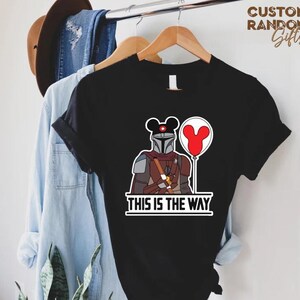 Puede incluir: Camiseta negra con una ilustraci&oacute;n de dibujos animados de un personaje mandaloriano, una cabeza de Mickey Mouse y un globo rojo. El texto "THIS IS THE WAY" est&aacute; impreso debajo. La camiseta est&aacute; colgada en una percha de madera.