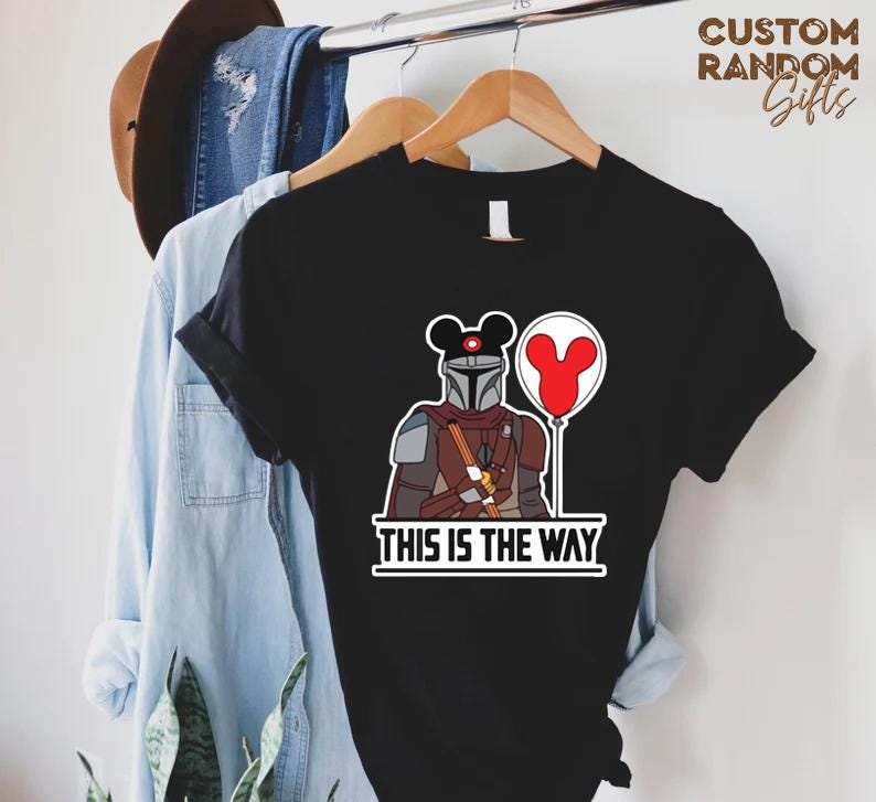 Puede incluir: Camiseta negra con una ilustraci&oacute;n de dibujos animados de un personaje mandaloriano, una cabeza de Mickey Mouse y un globo rojo. El texto "THIS IS THE WAY" est&aacute; impreso debajo. La camiseta est&aacute; colgada en una percha de madera.