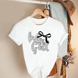 May include: White t-shirt with the text "but God" in a black, dotted font. The text is accompanied by a black bow and the phrase "He makes a way where there is no way". The shirt is hanging on a wooden hanger.