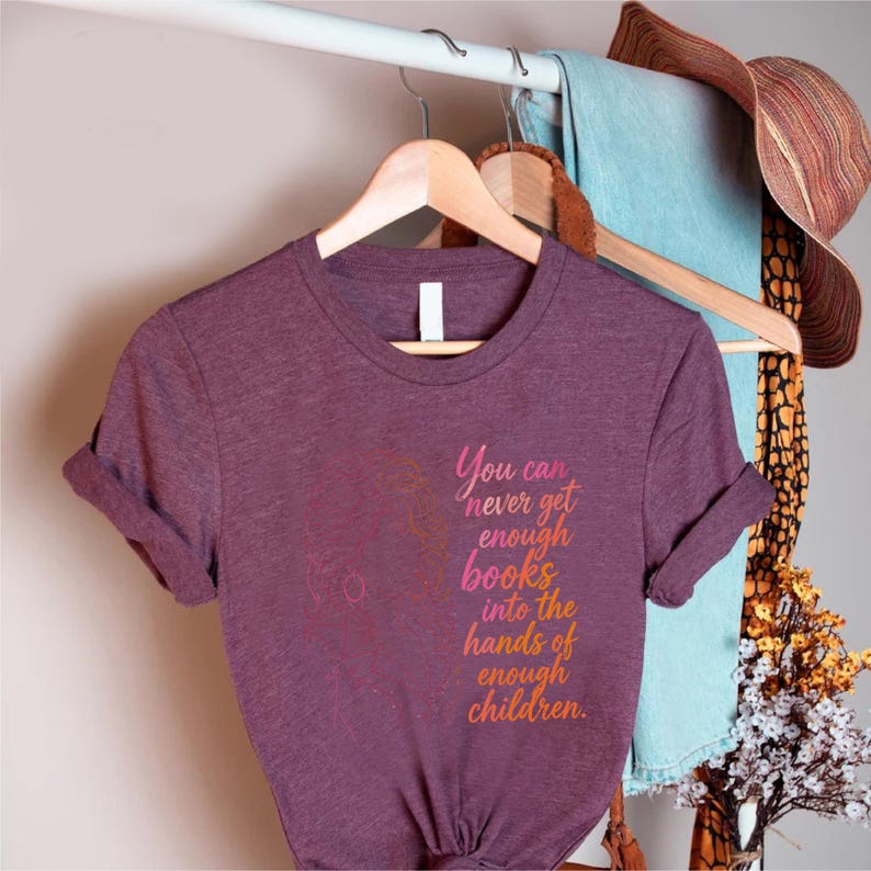 May include: A heathered plum short-sleeve t-shirt featuring a line art illustration of a person reading to a child. The text reads, You can never get enough books into the hands of enough children, in a gradient of pink, orange, and yellow.