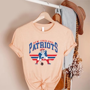 May include: A peach-colored t-shirt featuring the words "New England Patriots" in blue and red, with a vintage-style graphic of a Patriot football player. The shirt is hanging on a wooden hanger.