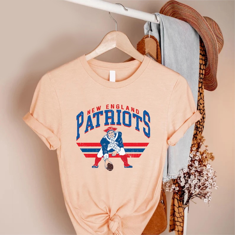 May include: A peach-colored t-shirt featuring the words "New England Patriots" in blue and red, with a vintage-style graphic of a Patriot football player. The shirt is hanging on a wooden hanger.