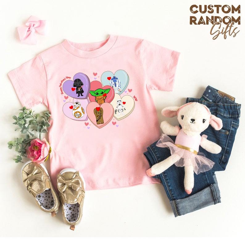 May include: A light pink t-shirt featuring Star Wars characters on heart-shaped candies. The shirt has the text "Happy Valentine's Day". Gold shoes, a denim pair of jeans, and a plush toy are also in the image.