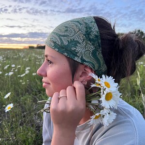 May include: A green paisley patterned headband is worn by a person in a field of daisies. The person is holding a small bouquet of daisies. The sky is a mix of blue, yellow, and orange.