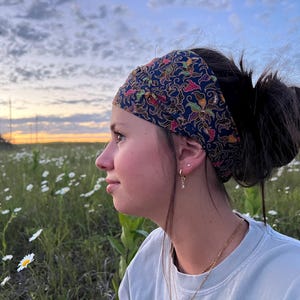 May include: A dark blue headband with a colorful floral pattern is worn by a person with dark hair. The person is in a field of white flowers, with a sunset in the background. The headband is the focal point of the image.