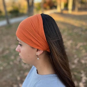 May include: An orange-colored headband made of textured fabric. The wide headband is worn across the forehead and covers the top of the head. The headband is paired with long, dark hair and a light blue shirt.