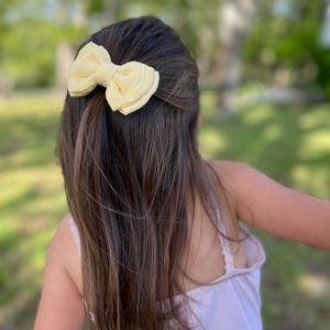 Könnte beinhalten: Eine hellgelbe Stoff-Haarspange mit Schleife. Die Schleife ist im klassischen Stil gebunden und am Haar befestigt. Das Haar ist lang und braun, mit Highlights. Der Hintergrund ist ein verschwommenes Grün, was auf eine Umgebung im Freien hindeutet.