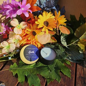 May include: A black candle in a round tin next to a candle lid with the text "Midnight Candles" and "Earth". The candles are surrounded by colorful flowers and green leaves on a wooden surface.