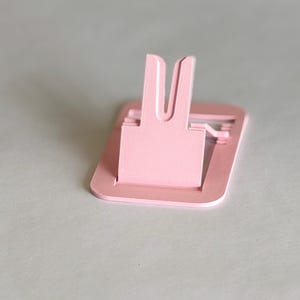 以下が含まれることがあります： 長方形のベースと上向きに回転するU字型のサポートアームを備えた、ライトピンクの折りたたみ式携帯電話スタンド。滑らかなマット仕上げの素材で作られており、モバイルデバイスを直立した状態で保持するように設計されています。