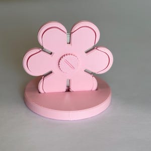 Può includere: Un supporto a forma di fiore rosa chiaro con una base circolare. L'elemento floreale presenta sei petali arrotondati e una manopola centrale circolare con una fessura diagonale, il tutto realizzato in materiale opaco con strati di stampa 3D.