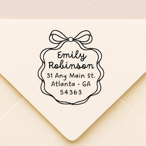 May include: A cream envelope with a black address stamp. The stamp has a bow design, the name "Emily Robinson", and the address: "31 Any Main St. Atlanta, GA 54363". The envelope is on a light background.