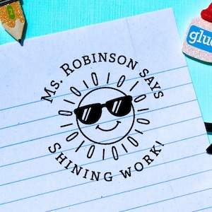 May include: A lined paper with a stamp that reads "Ms. Robinson says Shining Work!" with a smiling sun wearing sunglasses. A pencil, glue bottle, and scissors are also in the image.
