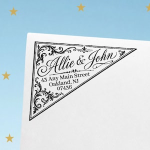 May include: A triangular black ink stamp impression on white paper featuring ornate filigree corner details and the text Allie & John, 43 Any Main Street, Oakland, NJ 07436. The design is framed by a thin black border with decorative flourishes.