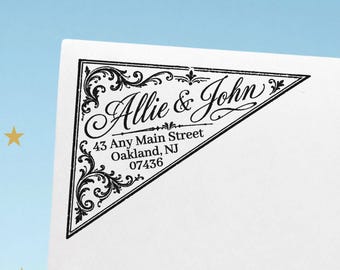 May include: A triangular black ink stamp impression on white paper featuring ornate filigree corner details and the text Allie & John, 43 Any Main Street, Oakland, NJ 07436. The design is framed by a thin black border with decorative flourishes.