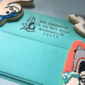May include: A turquoise blue envelope with a black and white shark graphic and the text "the Andersons 138 Any Main Street Manahawkin, NJ 08050" printed on the front.