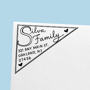 May include: A triangular black ink stamp impression on white paper featuring the text Silva Family, 321 Any Main St., Oakland, NJ 07436. The design is enclosed in a thin black triangular border with two small black hearts at the corners.
