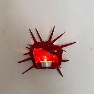 Pode incluir: Um suporte de vela de parede em cerâmica vermelha brilhante com formato de ouriço-do-mar ou estrela com espinhos. O centro oco contém uma pequena vela acesa, projetando um brilho quente nas paredes internas da estrutura vermelha.