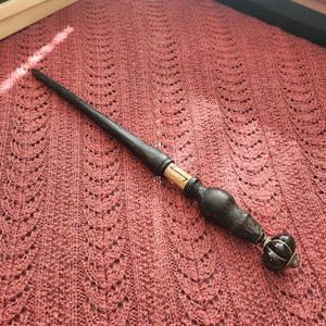 May include: A slender, dark-coloured wooden wand featuring a smooth, tapered shaft and a textured, bulbous handle. The handle includes a light-coloured section with a black rune symbol and terminates in a rounded, fluted, dark-coloured pommel cap.