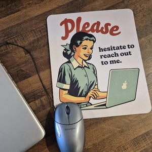 Puede incluir: Un ratón de ordenador y una alfombrilla de ratón con una ilustración retro de una mujer usando un portátil. La alfombrilla tiene el texto "Please hesitate to reach out to me" en rojo y negro.