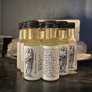Può includere: Una collezione di piccole bottiglie di vetro con tappi neri, riempite con un liquido giallo chiaro. Ogni bottiglia ha un'etichetta bianca con un'illustrazione dettagliata di un angelo e testo. Le bottiglie sono disposte su una superficie scura.