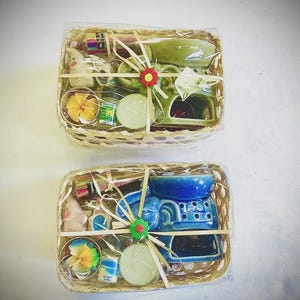 May include: Two rectangular woven baskets wrapped in clear plastic, each containing an elephant-shaped ceramic oil burner, a flower-shaped candle, a small bottle, and incense sticks. The top basket is green, and the bottom basket is blue.