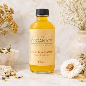 May include: A clear glass bottle filled with yellow liquid, labeled "BUTTER ME UP ORGANICS" and "Super Acne Fighter." The label also indicates the product contains Neem & Tea Tree and is 4 fl. oz. The bottle is set against a backdrop of flowers and a neutral background.