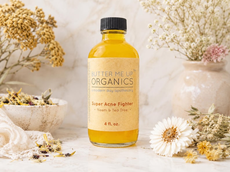 May include: A clear glass bottle filled with yellow liquid, labeled "BUTTER ME UP ORGANICS" and "Super Acne Fighter." The label also indicates the product contains Neem & Tea Tree and is 4 fl. oz. The bottle is set against a backdrop of flowers and a neutral background.