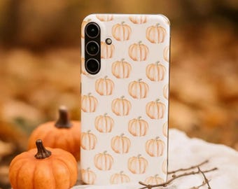 Puede incluir: Una funda de teléfono blanca con un patrón repetido de calabazas naranjas. La funda del teléfono está junto a dos calabazas pequeñas y una ramita. El fondo está borroso con colores otoñales.