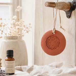 May include: A terracotta-colored, circular essential oil diffuser with an embossed mandala design hangs from a wooden peg. A small amber glass bottle of essential oil and a vase of dried flowers are in the background.