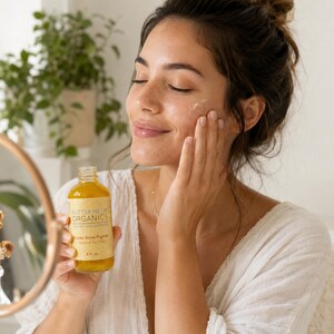 May include: A woman applying skincare product. She holds a yellow bottle of "BUTTER ME UP ORGANICS" serum, with the text "Super Active Fig Serum" visible. The bottle is 4 fl oz. The woman is wearing a white top, with a mirror and plants in the background.