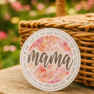 May include: A round sticker with the word "mama" in a cursive font, surrounded by a floral design in pink and white. The sticker has a white border with text that reads "Take care of the ones who raised you and those who love you."