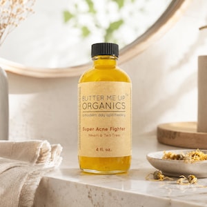 May include: A clear glass bottle with a black cap and a tan label that reads "BUTTER ME UP ORGANICS" and "Super Acne Fighter." The bottle contains a yellow liquid and is 4 fl. oz. A small ceramic dish with dried flowers sits nearby.