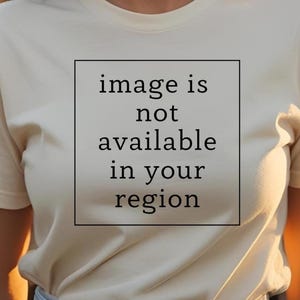 Könnte beinhalten: Cremefarbenes T-Shirt mit dem Text "image is not available in your region" in einem schwarzen Quadrat. Das Shirt hat kurze Ärmel und einen Rundhalsausschnitt.