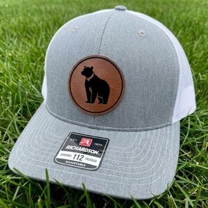 May include: Gray trucker hat with a white mesh back and a brown leather patch featuring a black bear silhouette. The hat has a curved brim and the text "RICHARDSON" and "ORIGINAL 112 TRUCKER" on a tag.