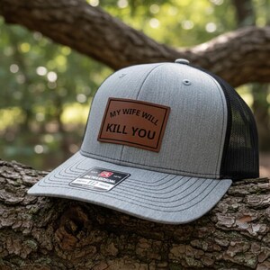 May include: Gray and black trucker hat with a brown leather patch that reads "MY WIFE WILL KILL YOU". The hat has a curved brim and a mesh back. The hat is sitting on a tree branch.