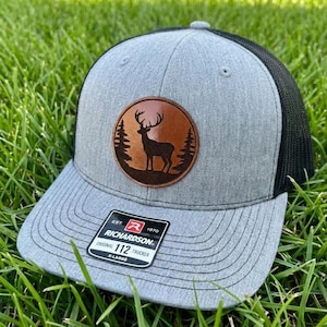 May include: A gray trucker hat with a black mesh back and a brown leather patch featuring a deer silhouette within a circle. The hat has a curved brim and the Richardson brand label.