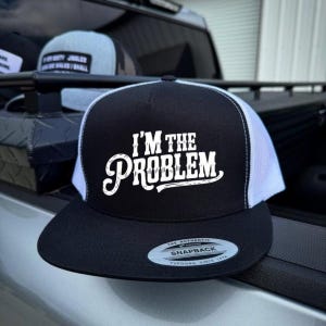 May include: Black and white snapback hat with the text "I'M THE PROBLEM" in white script. The hat has a black brim and a white mesh back. The hat is a Yupoong Snapback.