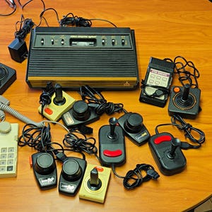 Könnte beinhalten: Eine schwarze Atari Video Computer System Konsole mit Holzmaserung, begleitet von mehreren kabelgebundenen Joysticks, Paddle-Controllern, einem Tastenfeld-Controller und einer Star Raiders-Auflage aus schwarzem, rotem und beigem Kunststoff.